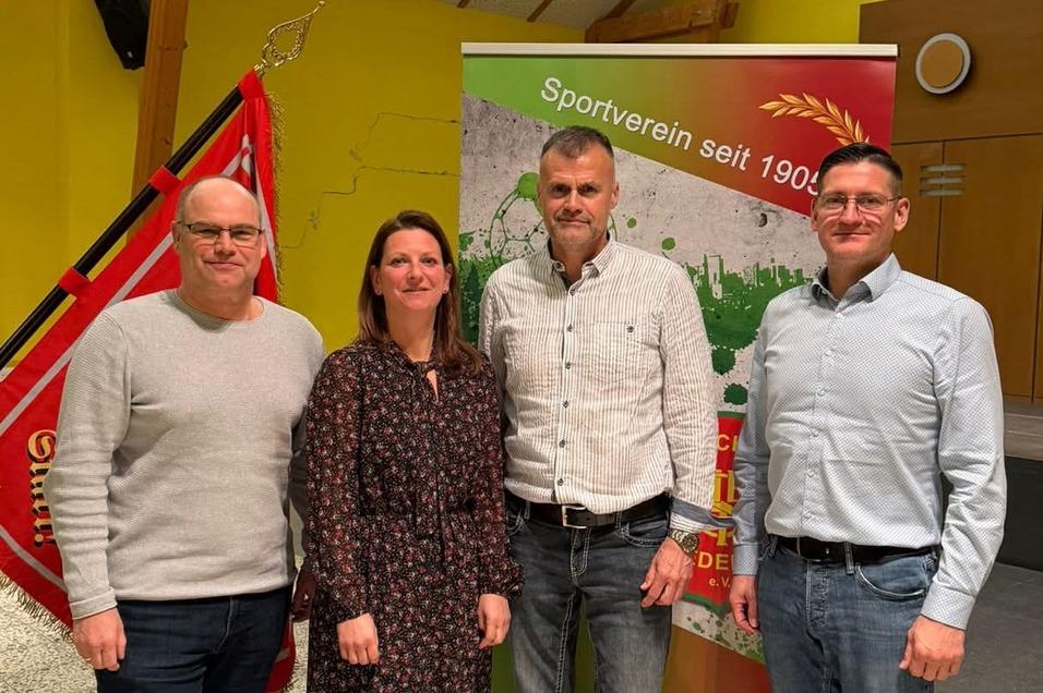 Vier Personen stehen lächelnd vor einem Banner des Sportvereins mit einer roten Fahne im Hintergrund, in einem hellen Raum.